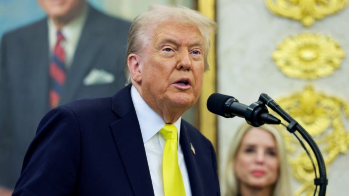 Donald Trump speaks at a microphone in the Oval Office, wearing a dark suit and yellow tie. A woman is blurred in the background.
