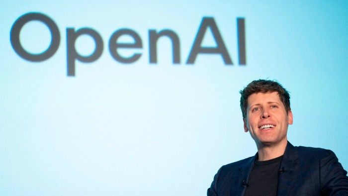 Sam Altman smiles while seated in front of a screen displaying the OpenAI logo.