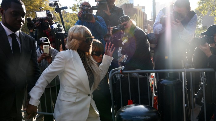 Janice Combs raises her hand while arriving at court, surrounded by security and photographers.