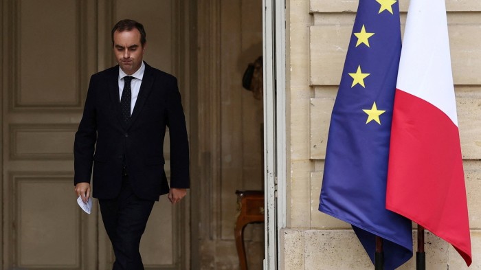 Sebastien Lecornu walks out of a doorway holding papers, near the EU and French flags, appearing solemn.