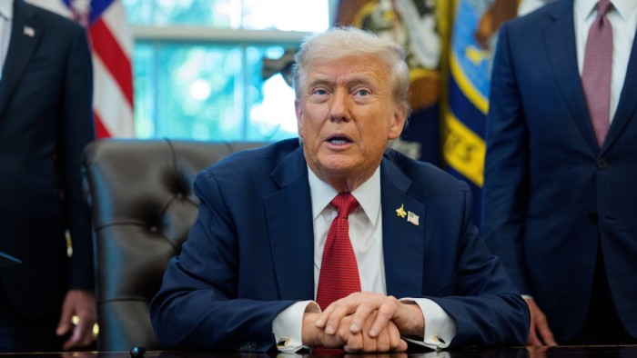Donald Trump sits at a desk in the Oval Office, speaking to the media after signing an executive order, with two men standing behind him