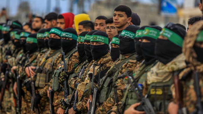 Fighters from the Ezz al-Din Al-Qassam brigades stand in a line holding weapons, wearing uniforms and green headbands.