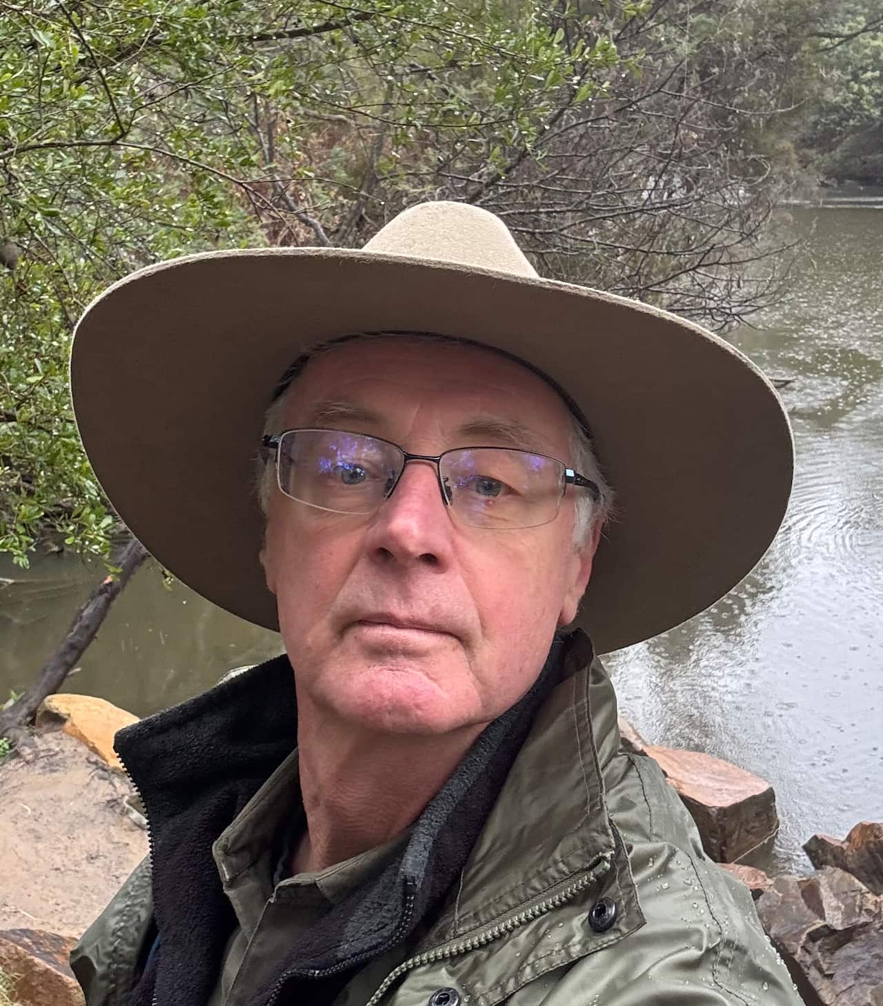 A man in a khaki jacket with a wide brimmed hate, standing in front of a river.