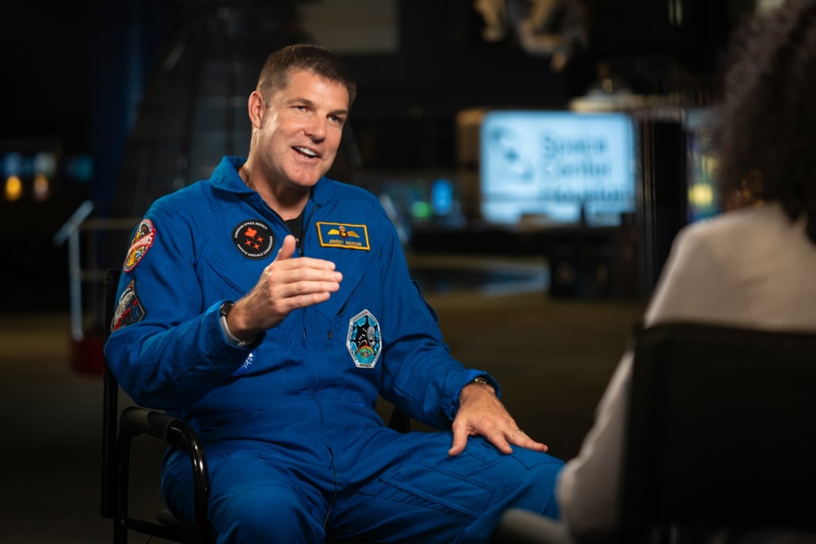 A man in an blue astronaut jumpsuit, raises his right arm as he speaks to a reporter.