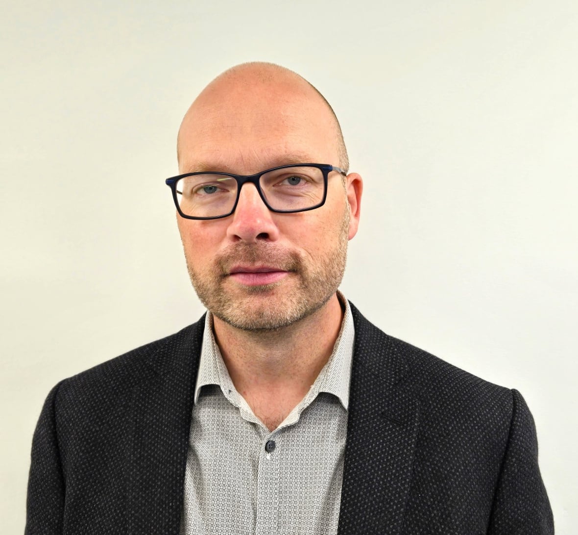 A man with black-rimmed glasses and a trim beard wears a dark suit jacket and a printed shirt.
