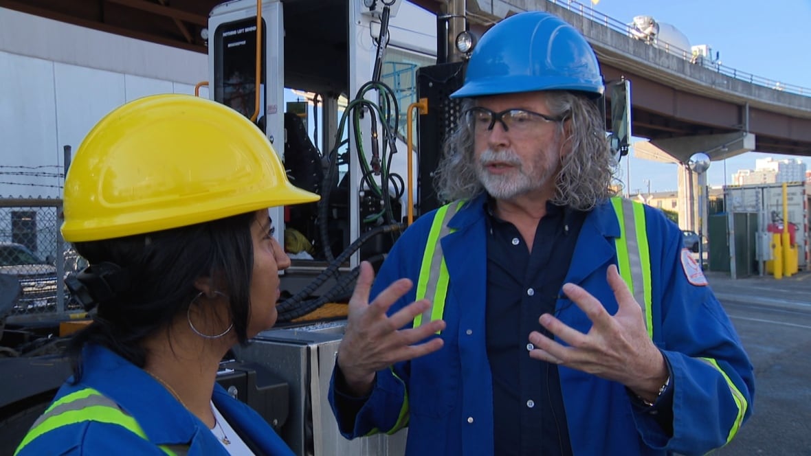A tall man with long, curly salt and pepper hair speaks with his hands outside in an industrial area. He wears a hard hat and protective glasses and a blue and yellow high viz vest. Beside him is a woman with dark hear wearing a similar outfit.