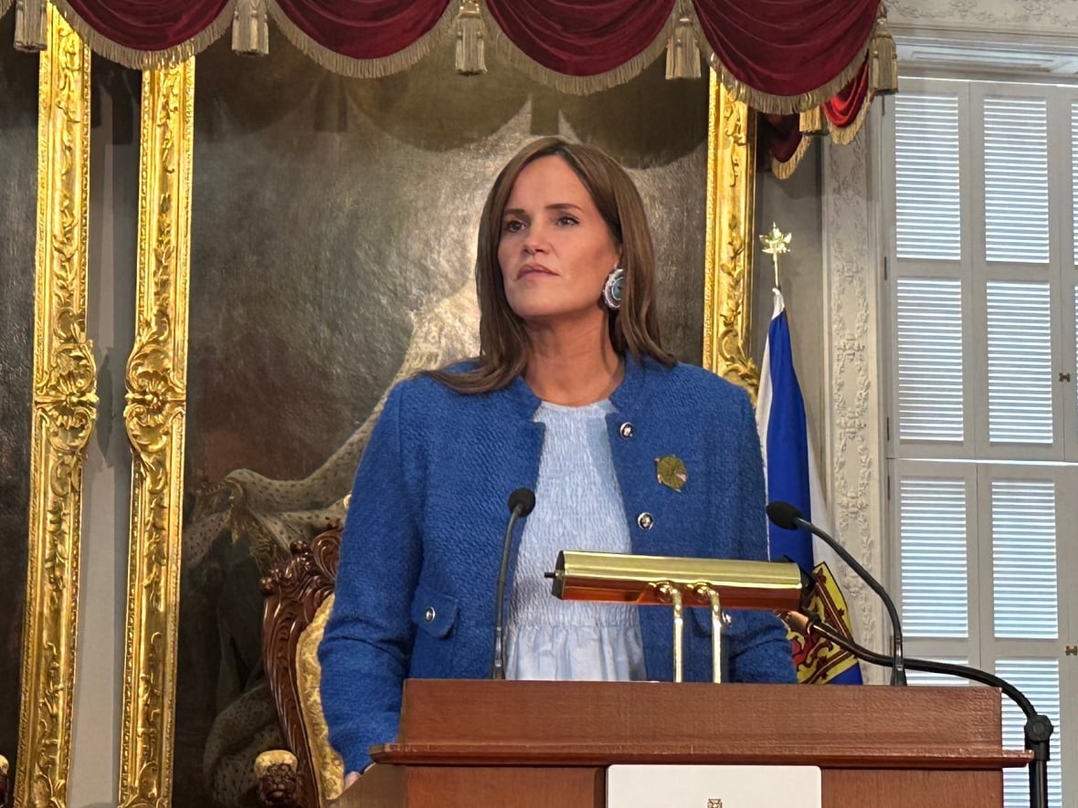 A woman in a blue blazer stands at a podium with ornately framed portraits hanging behind her.