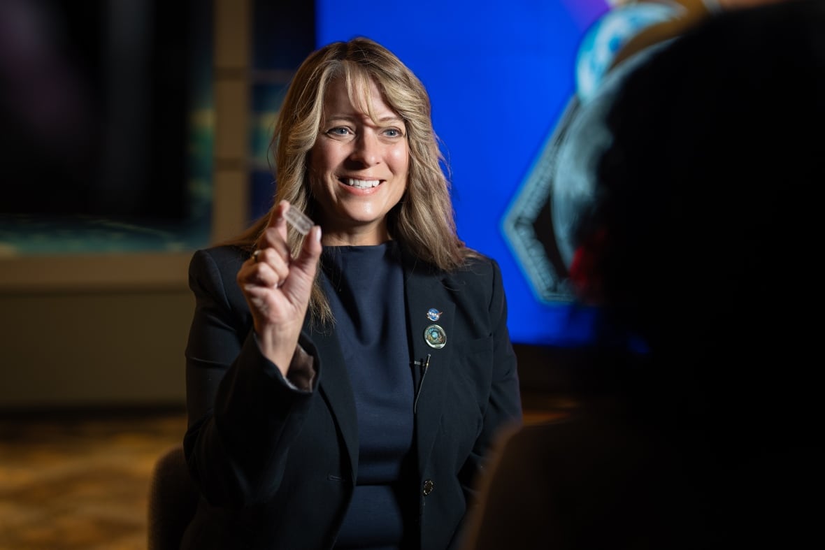 A woman with blonde hair in a black shirt and black jacket holds up a USB-sized chip.