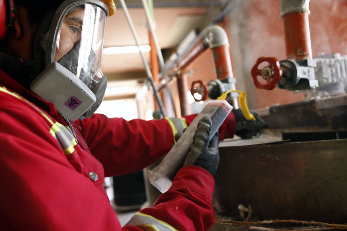 a worker has a gas mask on, protective gloves and a thick protective jacket. he's looking at a piece of technology in his hand, working in an industrial envrionment