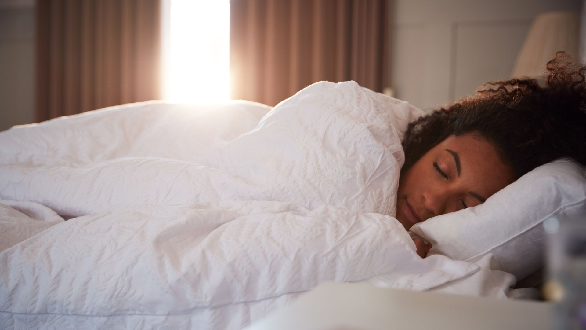 A woman sleeping soundly in bed under a white duvet, as early morning sun shines through the curtains behind her.