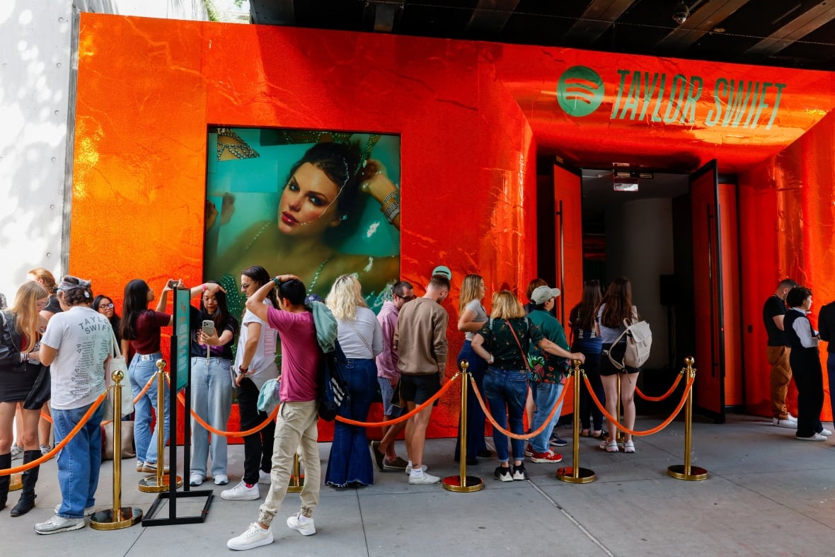 A group of people lined up on a sidewalk, behind a velvet rope, in front of a building with a bright orange facade, and the words "Taylor Swift" over the entrance. On the wall above the lined up people is an image of an album cover with a woman, Taylor Swift, laying in water with her hand behind her head. 
