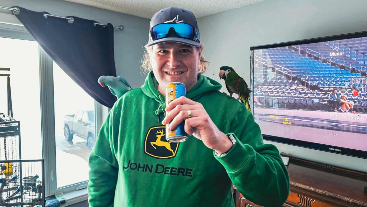 A man in a green sweater drinking out of a can with two parrots on his shoulders. 