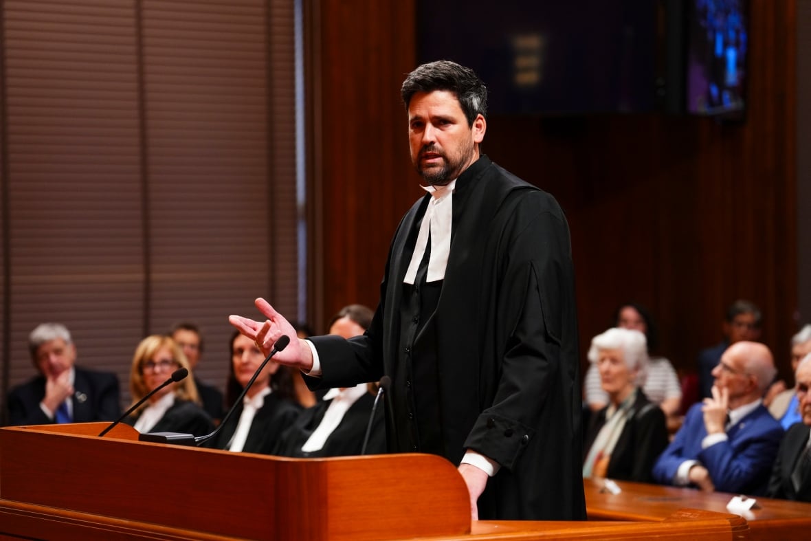 A man speaks at a podium, dressed in black legal robes.