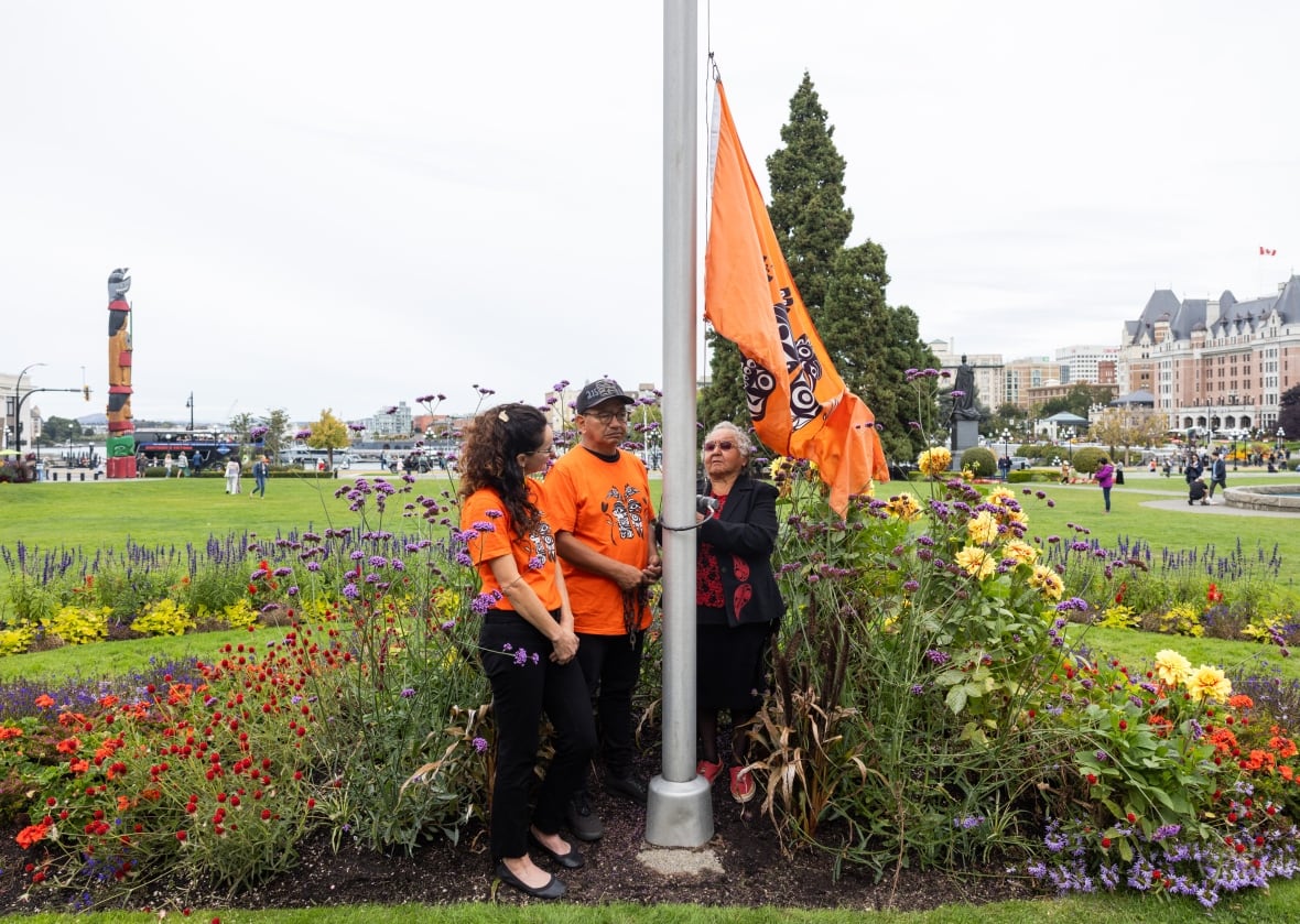 Three people standing beside a flagpole prepare to raise an orange flag.