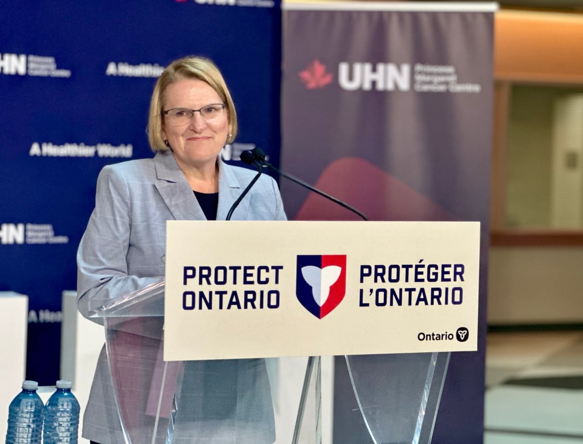 A woman with short, blonde hair and glasses stands in front of a podium with a sign reading Protect Ontario/Proteger L'Ontario. 