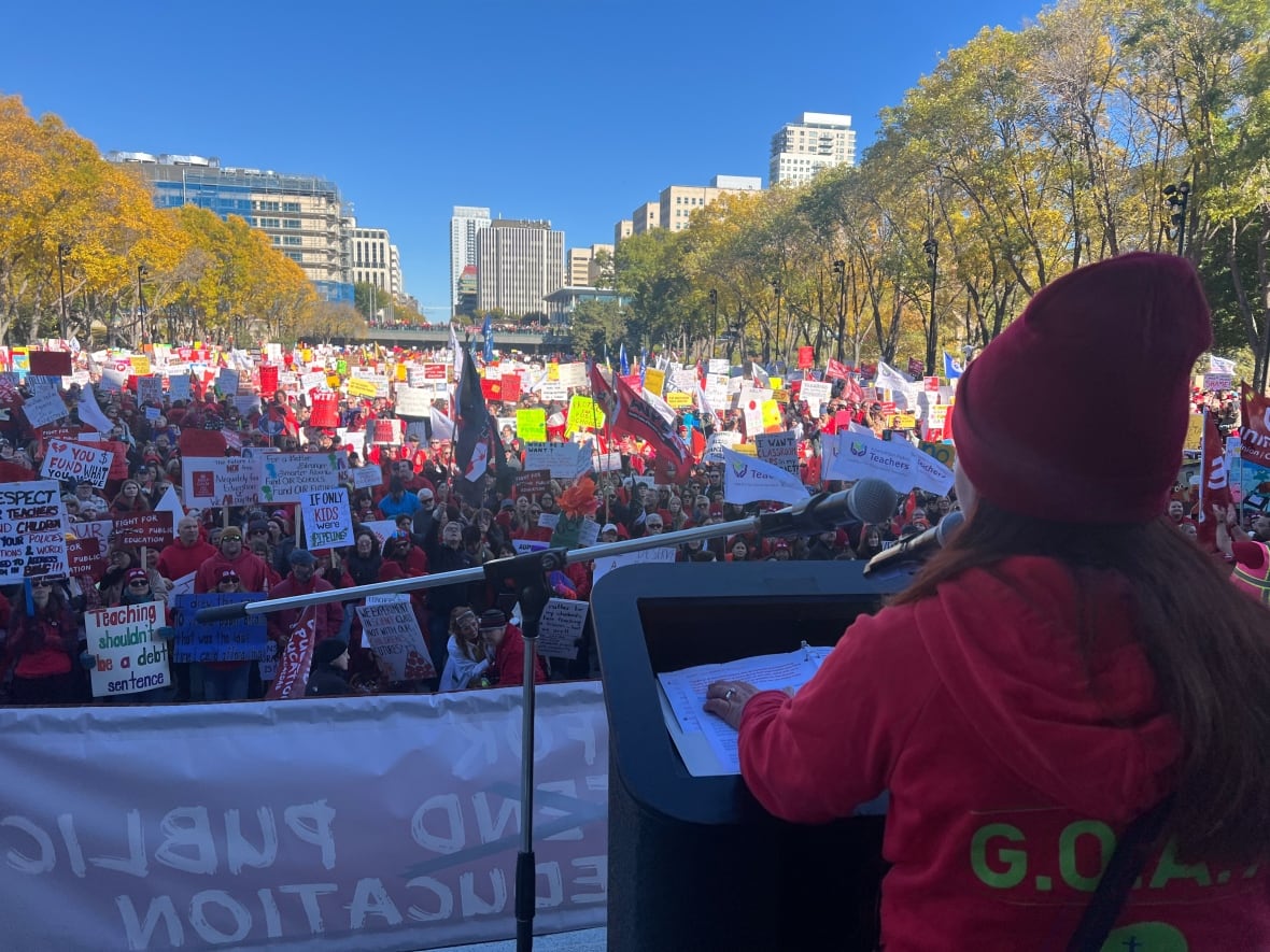A speaker at a podium looks onto a sea of people, who are holding signs and flags outdoors.