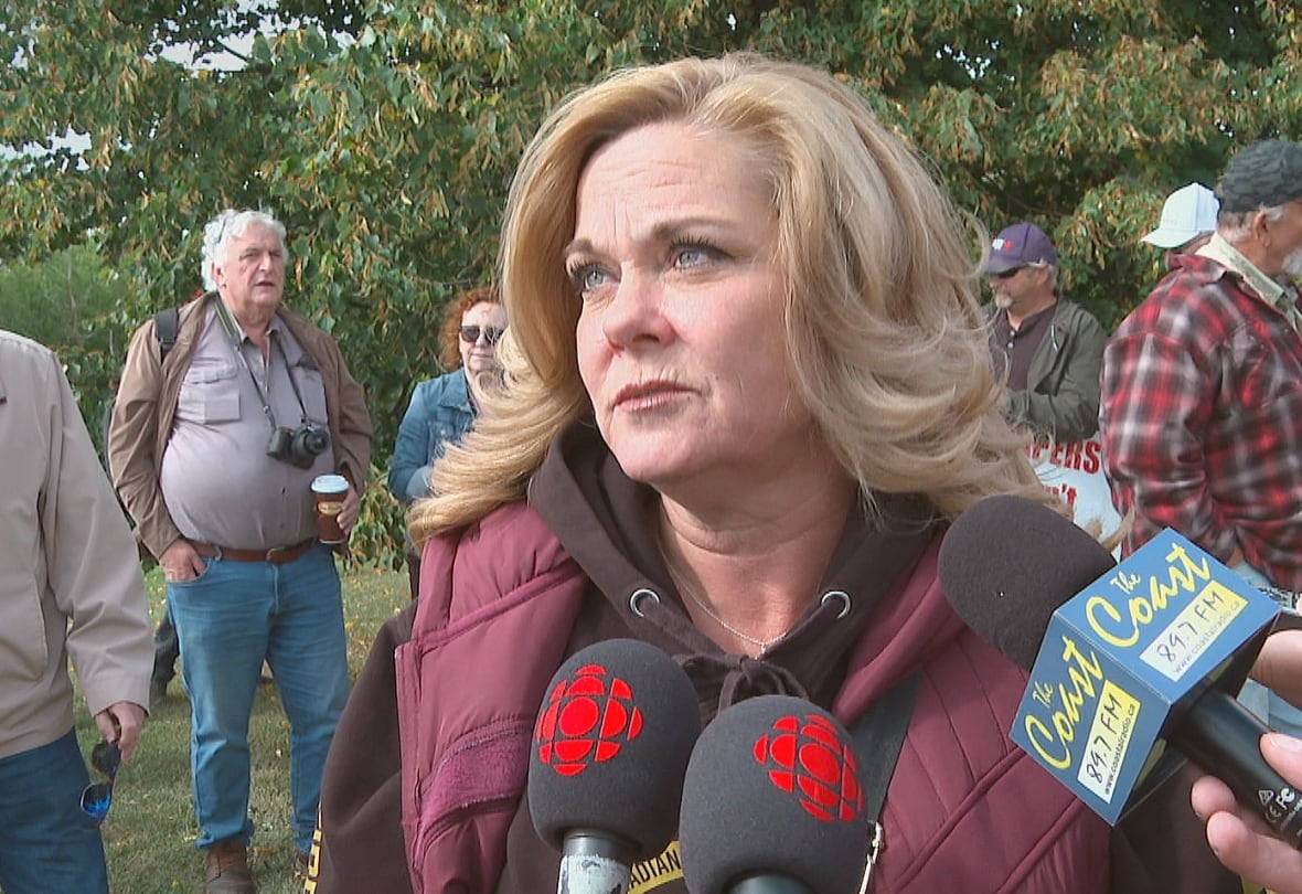 A woman with blonde hair, black hoodie and burgundy down filled vest speaks into several microphones.