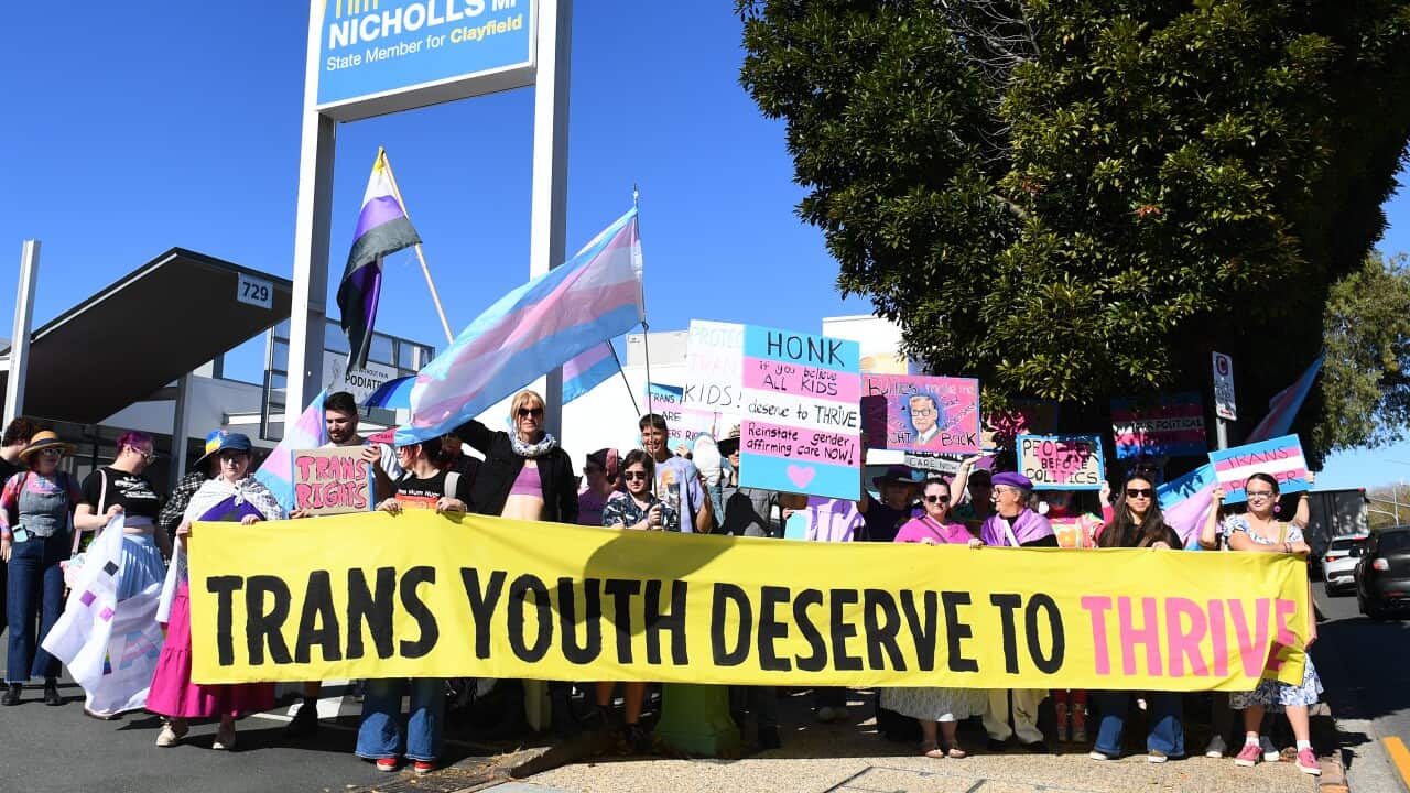 A group of transgender advocates protesting outside a politician's office, holding a yellow banner that reads "Trans youth deserve to thrive".