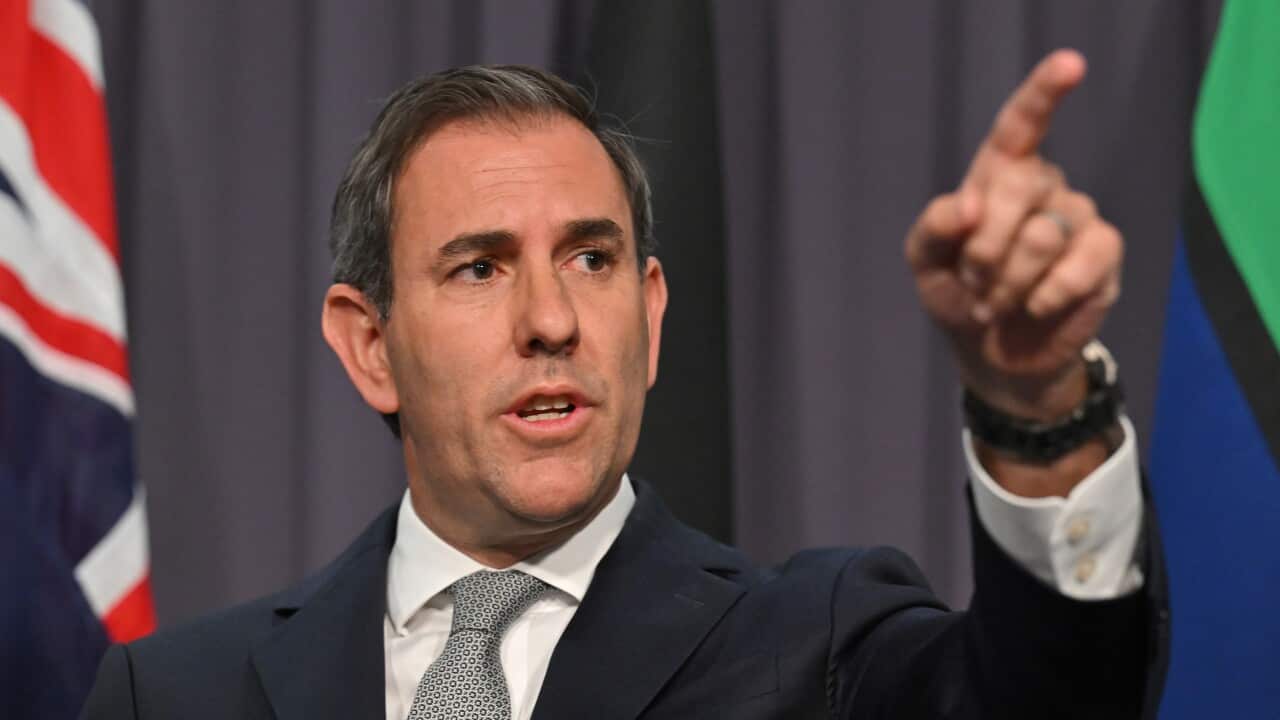 A man in a dark suit with a red tie, with the Australian and Aboriginal flags visible in the background.