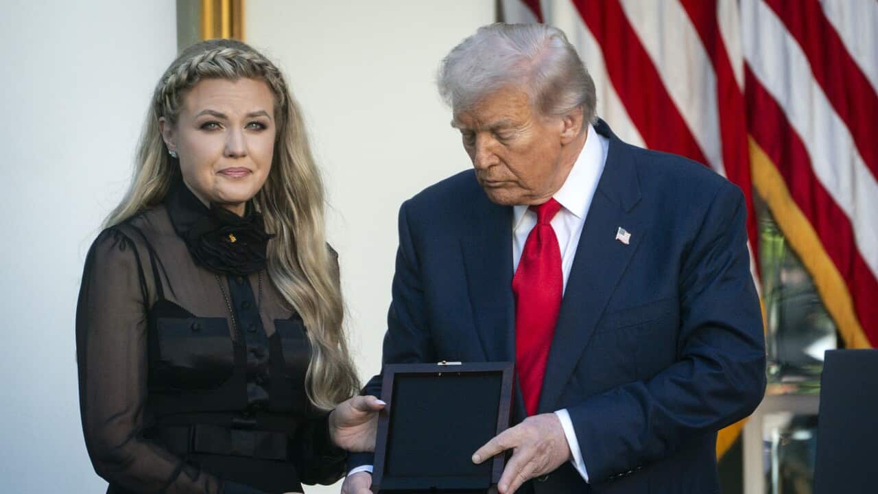 A woman in an all black outfit stands next to Donald Trump as he presents a medal to her with the American flag in the background.