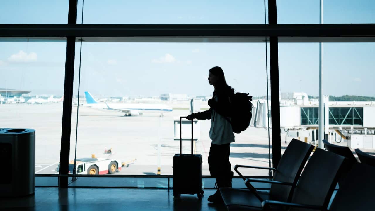 A woman with a suitcase standing in front of a large window at an airport, with planes and the tarmac visible behind her.
