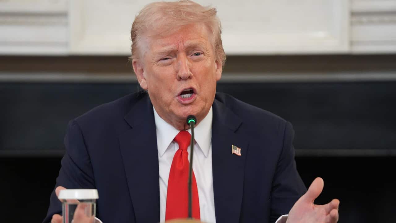 US President Donald Trump, wearing a suit and tie, is speaking in front of a microphone.