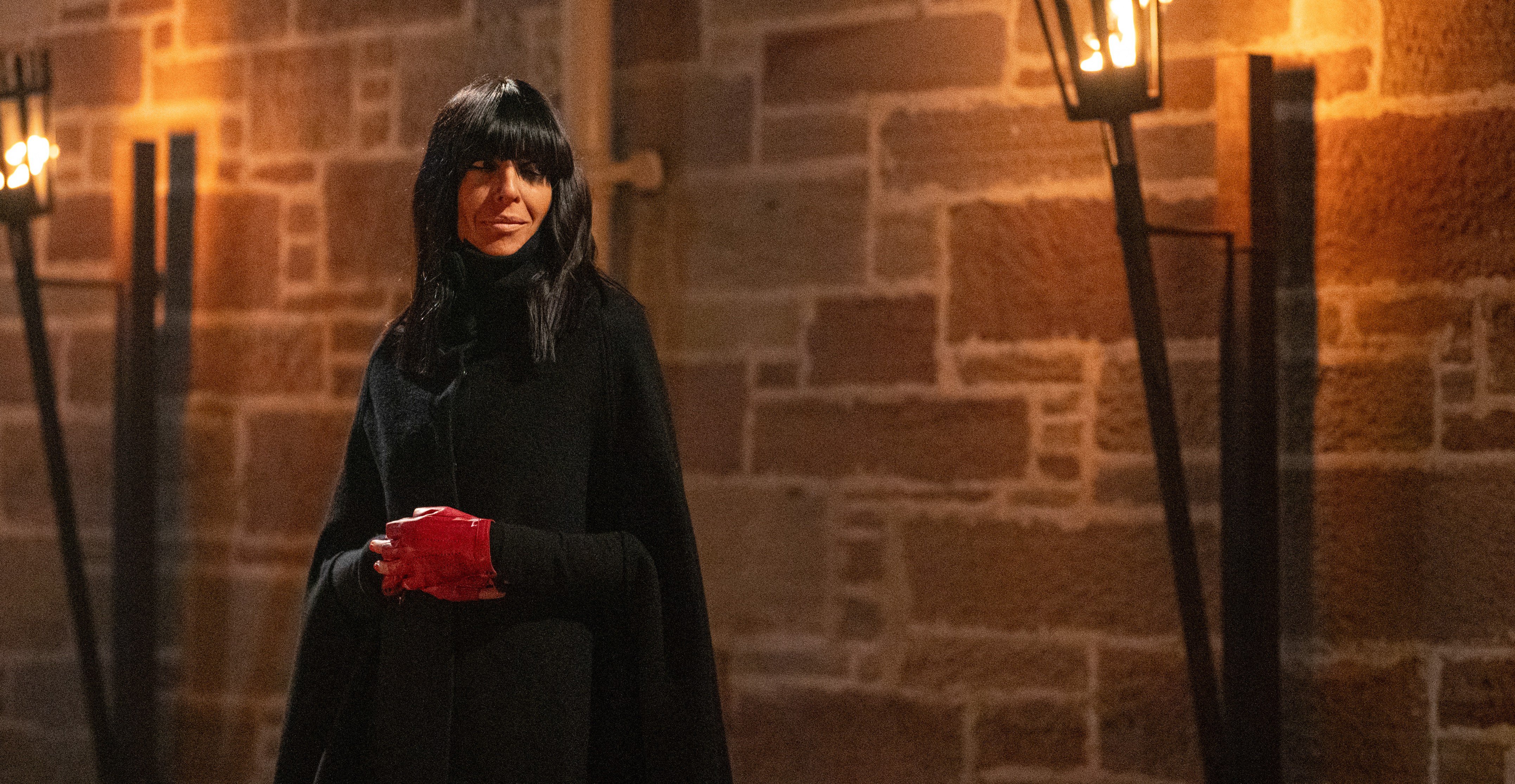 Claudia Winkleman in a black cape and red gloves, standing in front of a stone wall lit by two torches, promoting "The Celebrity Traitors".