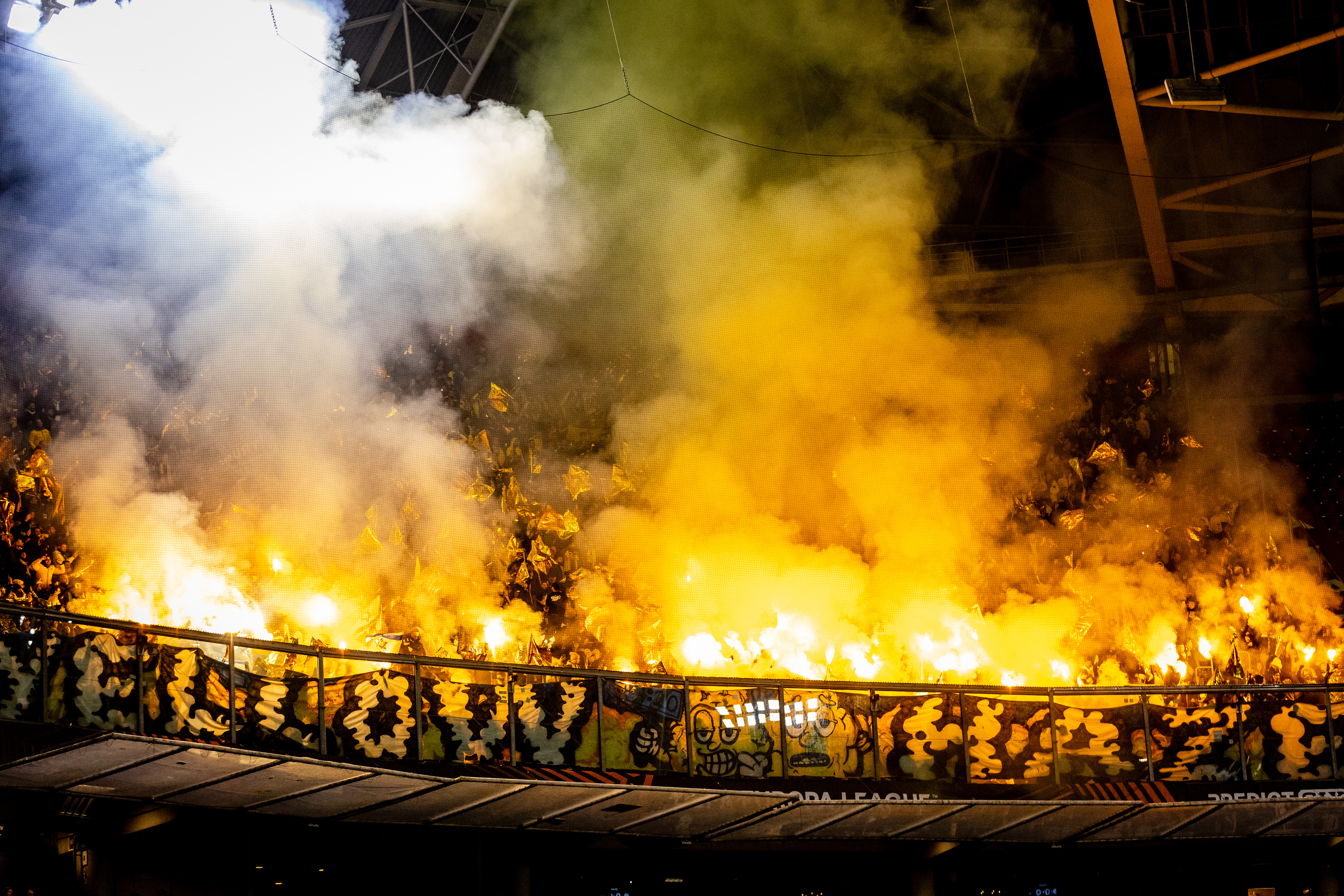 Fans of Maccabi Tel Aviv hold yellow flares, creating a wall of fire and smoke.