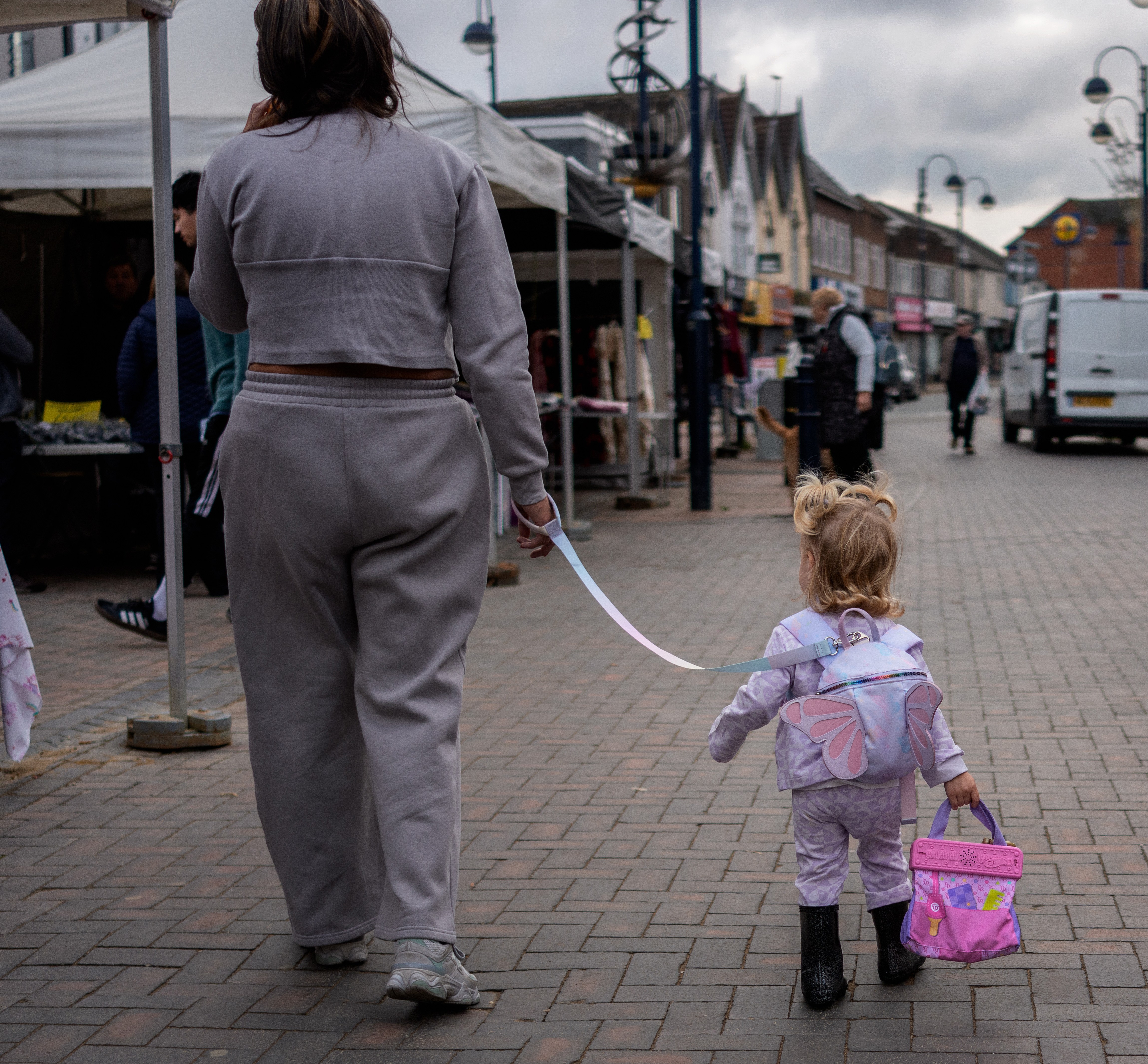 A woman holding a child's safety harness walks in front of the child on a street with shops and market stalls.