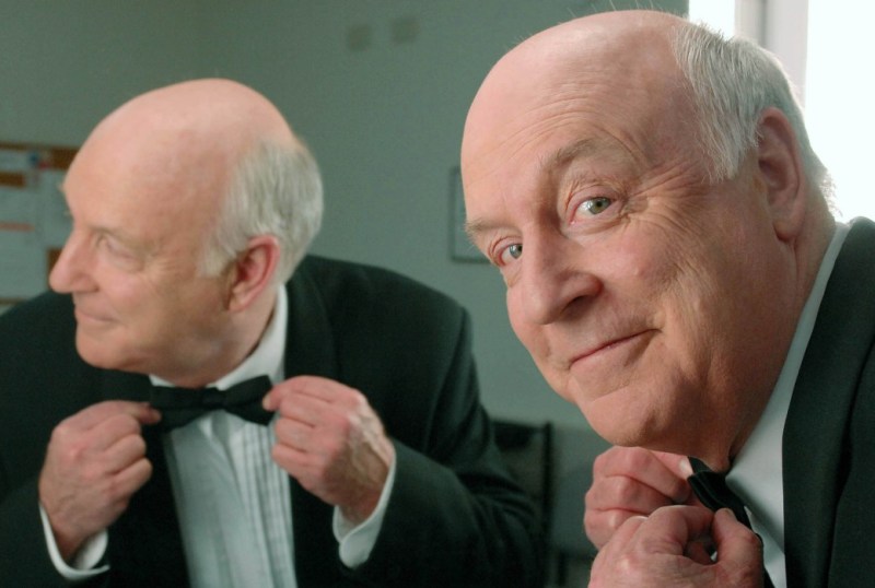 A mature man in a tuxedo adjusts his bow tie while looking into a mirror, smiling confidently. The setting appears to be a dressing room, with soft lighting and a neutral background.