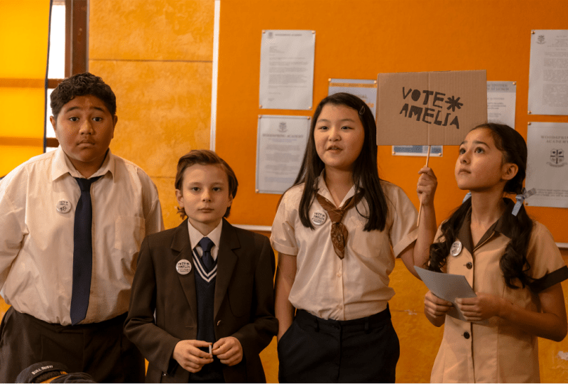 Group of schoolchildren participating in a mock election campaign, with one girl holding a sign that says "Vote Amelia." The students are wearing school uniforms and displaying campaign badges, set against a bright orange wall in a classroom environment.