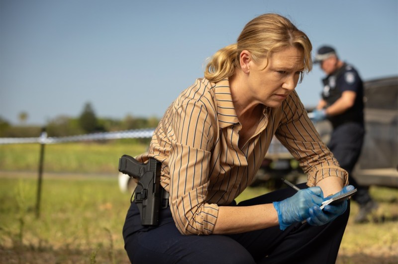 Police officer investigating a crime scene in rural Australia, wearing a striped shirt and blue gloves, crouched down while taking notes, with a firearm holstered at her side and a fellow officer in the background.
