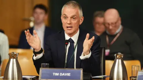 PA Media Tim Davie, sitting on a panel, wearing a suit and gesturing with his hands as he talks