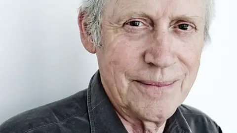 BBC Archie Fisher is looking into the camera and half smiling. He has grey hair but the photo has been cropped at his forehead. He is wearing a grey shirt.