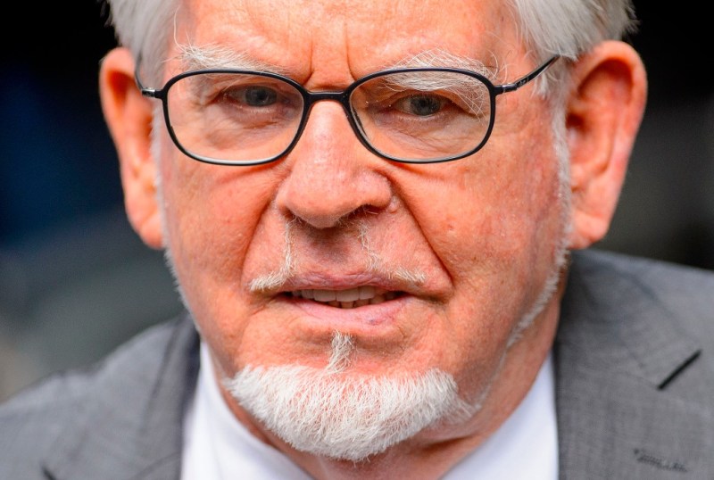 Close-up portrait of an elderly man with grey hair and glasses, featuring a distinctive white beard and moustache. He is wearing a grey suit, exuding a serious expression. The image captures the details of his facial features and attire, highlighting his age and wisdom.