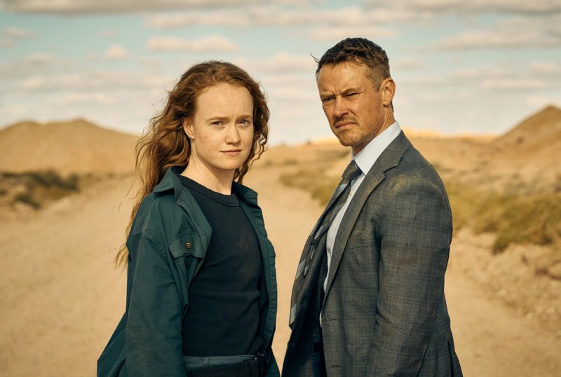 A woman in a green jacket and a man in a grey suit stand confidently on a dirt road in an Australian landscape, with sandy hills and a partly cloudy sky in the background.