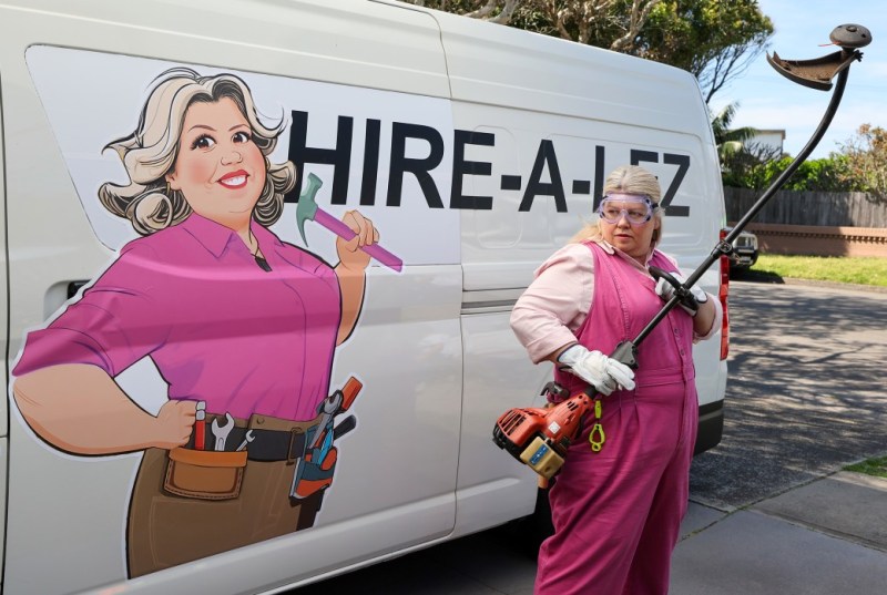 A woman in pink overalls and safety goggles stands beside a van featuring a cartoon image of herself, holding a garden tool. The van displays the business name "HIRE-A-LEZ," indicating a service for landscaping or home repairs.