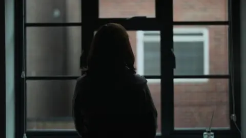 A woman, completely in shadow, stands with her back to the camera and looks out of a window onto a brick wall.