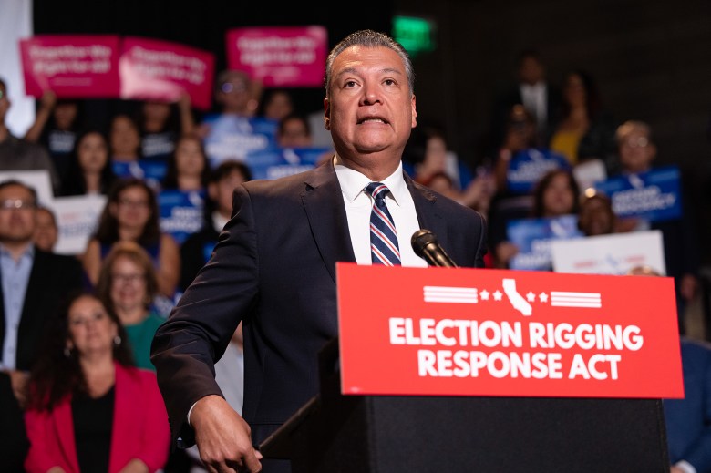 A person, wearing a black suit with a stripped-pattern tie, stands in front of a podium with a red sign on the front that says, "elections rigging response act" during a press conference. A crowd of attendees, many holding signs, can be seen standing in the background.