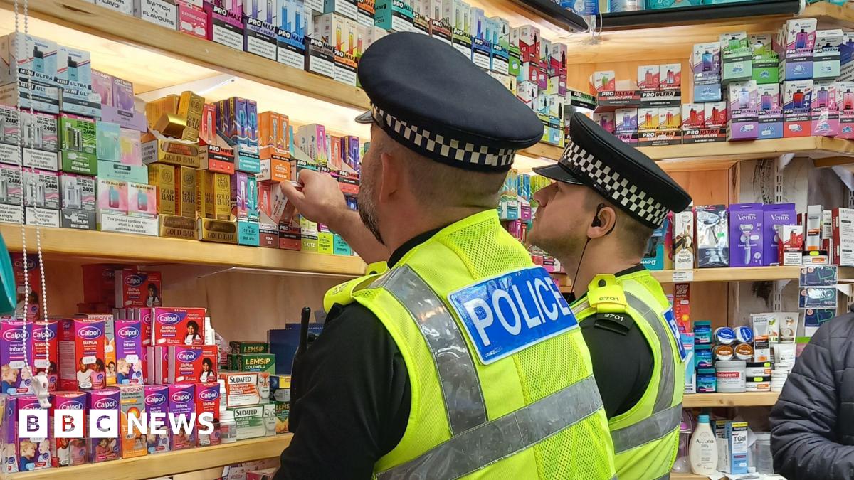 Two male police officers, wearing police caps and high-vis police branded vests, examine vapes on a shelf at a store.