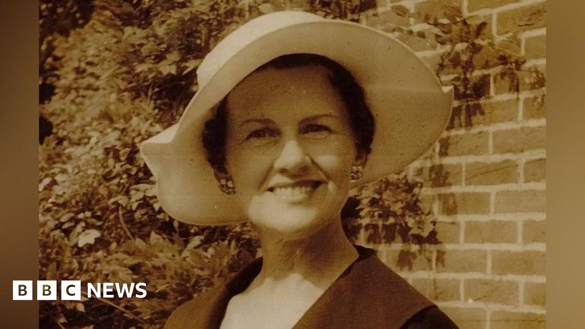 A headshot photo of Muriel McKay standing outdoors with a wall behind her that is partially covered in the creeper wisteria. She wears a flattering white rimmed hat, cluster earrings and a dark coloured coat. The photo is sepia coloured.