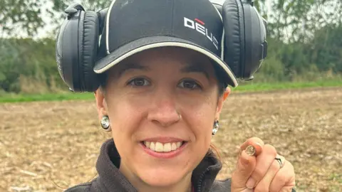 Emma Youell A smiling woman holds up a tiny coin to the camera in a head and shoulders shot. She's wearing a dark fleece, black baseball cap and large black headphones