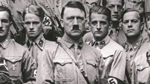 Getty Images Adolf Hitler staring at camera next to young unsmiling men in Nazi uniform in a black and white photo.