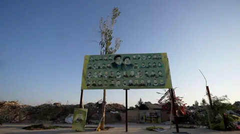 Neha Sharma/BBC A large green billboard on stilts displays images of men on a green background. Behind the billboard is rubble and destroyed buildings.