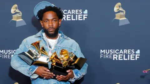 Reuters Kendrick Lamar wearing a denim jacket and light denim cap holds four grammys in his hands