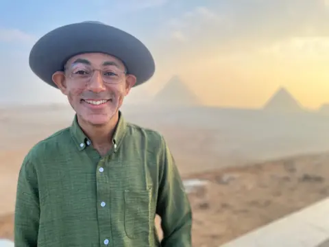 Ahmed Seddik smiles straight down the camera. He wears a green shirt, glasses and a large hat. In the background are two of the Pyramids of Giza. 