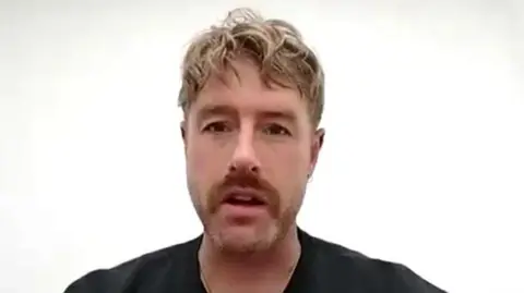 Christian Aldridge talks during a Zoom call. He is sat in front of a white wall. He has short but wavy blonde hair and has a stubbly beard. He is wearing a black t-shirt.