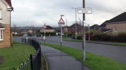 BBC Road approaching a roundabout with modern houses on either side. There is a sign saying Brambling Road as well as one depicting speed humps and another a roundabout.