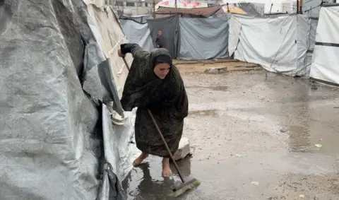 A Palestinian woman brushes away water on the ground in front of a tent