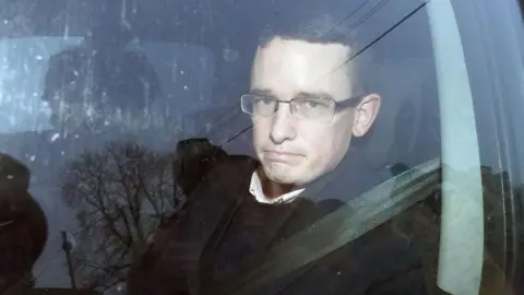 PA Enoch Burke is sitting in a car looking out. He's wearing a seat belt. He has glasses on. 